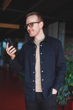Optik gözlüklü mutlu bir adamın yarım boy portresi modern cep telefonunu tutuyor ve kameraya gülümsüyor, dijital akıllı telefonlu neşeli blogcu birlikte çalışma alanında poz veriyor.