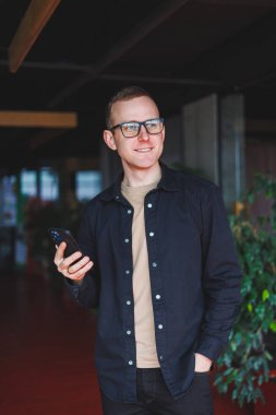 Optik gözlüklü mutlu bir adamın yarım boy portresi modern cep telefonunu tutuyor ve kameraya gülümsüyor, dijital akıllı telefonlu neşeli blogcu birlikte çalışma alanında poz veriyor.