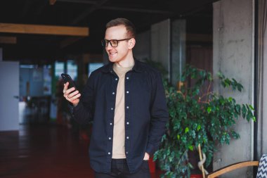 Optik gözlüklü mutlu bir adamın yarım boy portresi modern cep telefonunu tutuyor ve kameraya gülümsüyor, dijital akıllı telefonlu neşeli blogcu birlikte çalışma alanında poz veriyor.