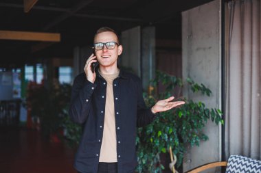 Optik gözlüklü mutlu bir adamın yarım boy portresi modern cep telefonunu tutuyor ve kameraya gülümsüyor, dijital akıllı telefonlu neşeli blogcu birlikte çalışma alanında poz veriyor.