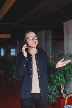 Optik gözlüklü mutlu bir adamın yarım boy portresi modern cep telefonunu tutuyor ve kameraya gülümsüyor, dijital akıllı telefonlu neşeli blogcu birlikte çalışma alanında poz veriyor.