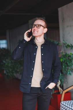 Half-length portrait of happy man in optical glasses holding modern mobile phone and smiling at camera, cheerful blogger with digital smartphone posing in coworking space