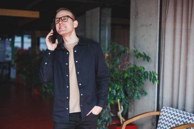 Optik gözlüklü mutlu bir adamın yarım boy portresi modern cep telefonunu tutuyor ve kameraya gülümsüyor, dijital akıllı telefonlu neşeli blogcu birlikte çalışma alanında poz veriyor.