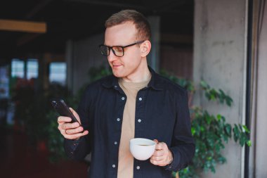 Elinde modern bir cep telefonu ve kahve tutan, optik gözlüklü mutlu bir adamın portresi, kafede dijital akıllı telefonuyla poz veren neşeli bir blogcu.