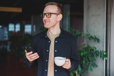 Elinde modern bir cep telefonu ve kahve tutan, optik gözlüklü mutlu bir adamın portresi, kafede dijital akıllı telefonuyla poz veren neşeli bir blogcu.