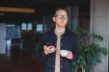 Elinde modern bir cep telefonu ve kahve tutan, optik gözlüklü mutlu bir adamın portresi, kafede dijital akıllı telefonuyla poz veren neşeli bir blogcu.