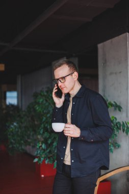 Elinde modern bir cep telefonu ve kahve tutan, optik gözlüklü mutlu bir adamın portresi, kafede dijital akıllı telefonuyla poz veren neşeli bir blogcu.