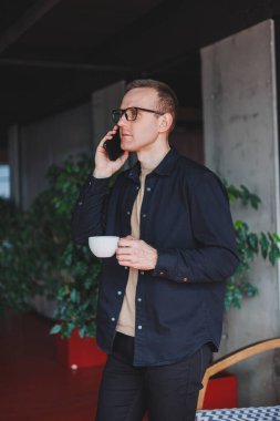 Elinde modern bir cep telefonu ve kahve tutan, optik gözlüklü mutlu bir adamın portresi, kafede dijital akıllı telefonuyla poz veren neşeli bir blogcu.