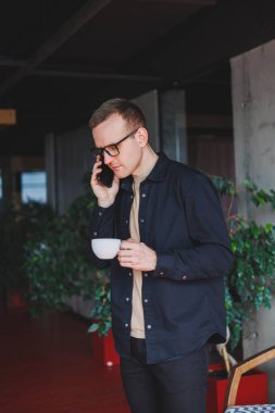 Portrait of a happy successful man in optical glasses holding a modern mobile phone and coffee in his hands, a cheerful blogger with a digital smartphone posing in a cafe