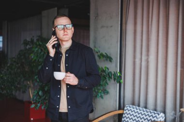 Elinde modern bir cep telefonu ve kahve tutan, optik gözlüklü mutlu bir adamın portresi, kafede dijital akıllı telefonuyla poz veren neşeli bir blogcu.