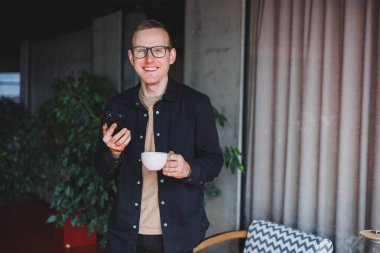 Elinde modern bir cep telefonu ve kahve tutan, optik gözlüklü mutlu bir adamın portresi, kafede dijital akıllı telefonuyla poz veren neşeli bir blogcu.