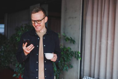 Elinde modern bir cep telefonu ve kahve tutan, optik gözlüklü mutlu bir adamın portresi, kafede dijital akıllı telefonuyla poz veren neşeli bir blogcu.