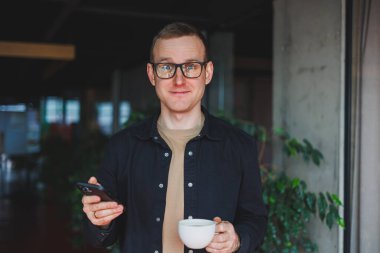 Elinde modern bir cep telefonu ve kahve tutan, optik gözlüklü mutlu bir adamın portresi, kafede dijital akıllı telefonuyla poz veren neşeli bir blogcu.