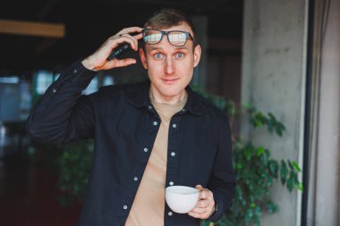 Elinde modern bir cep telefonu ve kahve tutan, optik gözlüklü mutlu bir adamın portresi, kafede dijital akıllı telefonuyla poz veren neşeli bir blogcu.