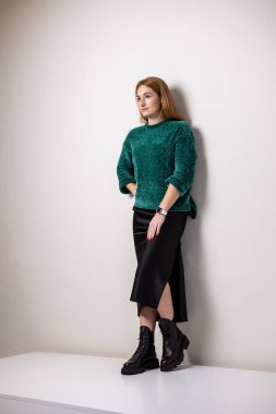 A woman in a green warm sweater and a black skirt in black leather boots. Fashionable autumn outfit.