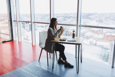 Modern çalışma ortamında açık bir ağ kitabıyla oturan ve günlük kıyafetlerle cep telefonundan bildirileri kontrol eden kadın serbest çalışan..
