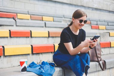 Şehrin dışında oturan, akıllı telefonuyla başka tarafa bakan ve gülümseyen rahat genç bir kadının portresi.