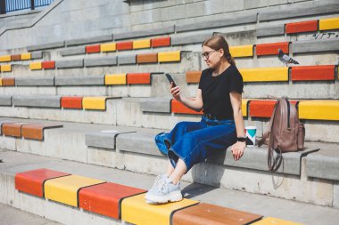 Şehrin dışında oturan, akıllı telefonuyla başka tarafa bakan ve gülümseyen rahat genç bir kadının portresi.