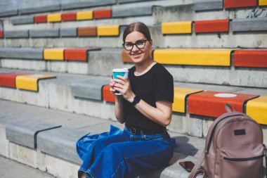 Kadın merdivenlerde oturuyor ve elinde tek kullanımlık bir fincan kahve tutuyor..