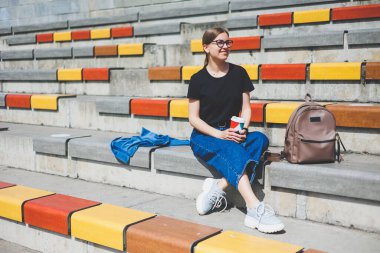 Kadın merdivenlerde oturuyor ve elinde tek kullanımlık bir fincan kahve tutuyor..