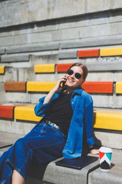 Genç ve şık bir iş kadını merdivenlerde oturuyor ve elinde telefon ve bir fincan kahve tutuyor.