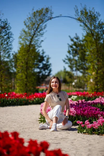 Back view of anonymous woman in summer dress and hat walking near blooming tulips in meadow on sunny day in nature