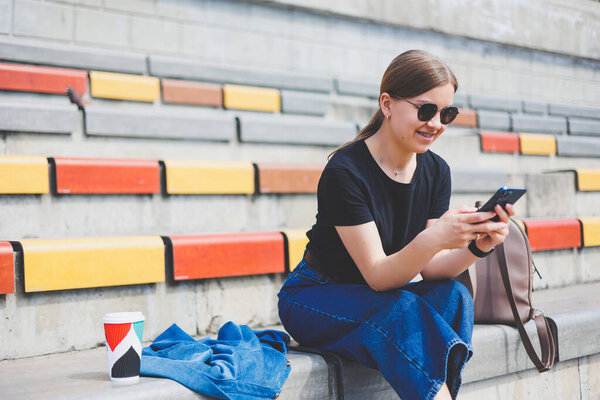 Portrait of relaxed young woman sitting outside in the city with her smart phone looking away and smiling