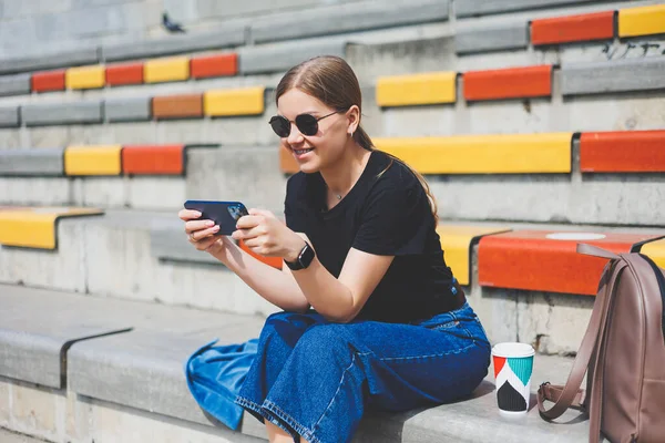 Şehrin dışında oturan, akıllı telefonuyla başka tarafa bakan ve gülümseyen rahat genç bir kadının portresi.