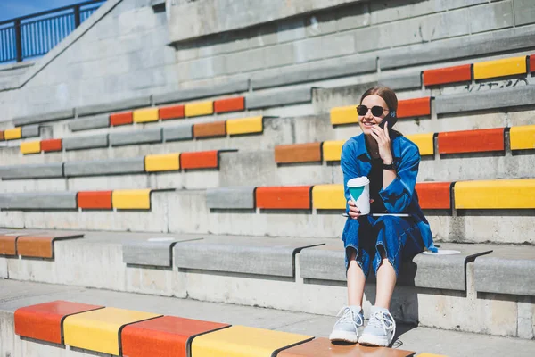 Kadın merdivenlerde oturuyor ve elinde tek kullanımlık bir fincan kahve tutuyor..