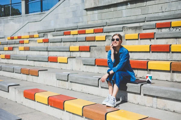 Genç ve şık bir iş kadını merdivenlerde oturuyor ve elinde telefon ve bir fincan kahve tutuyor.