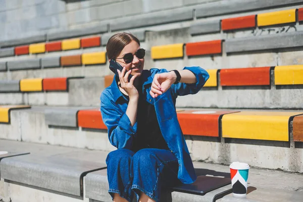 Genç ve şık bir iş kadını merdivenlerde oturuyor ve elinde telefon ve bir fincan kahve tutuyor.