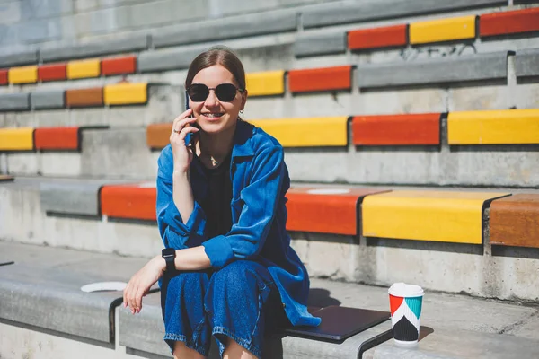 Genç ve şık bir iş kadını merdivenlerde oturuyor ve elinde telefon ve bir fincan kahve tutuyor.