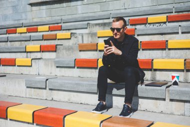 Şehirdeki şehir merkezinde dinlenirken gülümseyen modaya uygun giyinmiş neşeli hippi adam boş zamanlarında cep telefonu kullanıyor. Güneş gözlüklü mutlu Orta Doğulu erkek blogcu seyahat yaşam tarzının keyfini çıkarıyor.