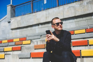 Şehirdeki şehir merkezinde dinlenirken gülümseyen modaya uygun giyinmiş neşeli hippi adam boş zamanlarında cep telefonu kullanıyor. Güneş gözlüklü mutlu Orta Doğulu erkek blogcu seyahat yaşam tarzının keyfini çıkarıyor.