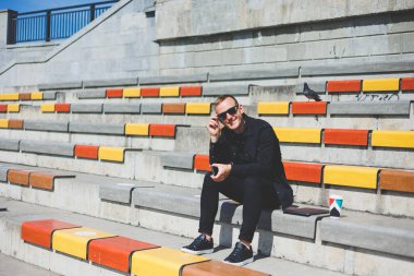 Şehirdeki şehir merkezinde dinlenirken gülümseyen modaya uygun giyinmiş neşeli hippi adam boş zamanlarında cep telefonu kullanıyor. Güneş gözlüklü mutlu Orta Doğulu erkek blogcu seyahat yaşam tarzının keyfini çıkarıyor.