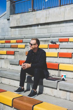 Şehirdeki şehir merkezinde dinlenirken gülümseyen modaya uygun giyinmiş neşeli hippi adam boş zamanlarında cep telefonu kullanıyor. Güneş gözlüklü mutlu Orta Doğulu erkek blogcu seyahat yaşam tarzının keyfini çıkarıyor.