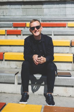 Şehirdeki şehir merkezinde dinlenirken gülümseyen modaya uygun giyinmiş neşeli hippi adam boş zamanlarında cep telefonu kullanıyor. Güneş gözlüklü mutlu Orta Doğulu erkek blogcu seyahat yaşam tarzının keyfini çıkarıyor.