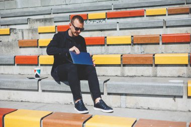 Genç işadamı ofisinin önündeki bankta otururken bilgisayarla çalışıyor.