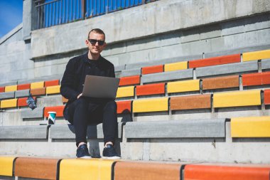 Genç işadamı ofisinin önündeki bankta otururken bilgisayarla çalışıyor.