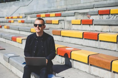 Genç işadamı ofisinin önündeki bankta otururken bilgisayarla çalışıyor.