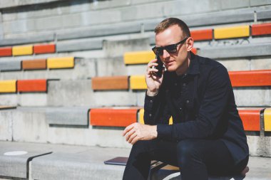 Şehirdeki şehir merkezinde dinlenirken gülümseyen modaya uygun giyinmiş neşeli hippi adam boş zamanlarında cep telefonu kullanıyor. Güneş gözlüklü mutlu Orta Doğulu erkek blogcu seyahat yaşam tarzının keyfini çıkarıyor.