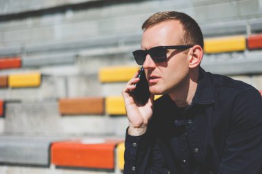 Şehirdeki şehir merkezinde dinlenirken gülümseyen modaya uygun giyinmiş neşeli hippi adam boş zamanlarında cep telefonu kullanıyor. Güneş gözlüklü mutlu Orta Doğulu erkek blogcu seyahat yaşam tarzının keyfini çıkarıyor.