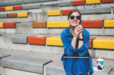 Modern Park Caddesi 'nde dizüstü bilgisayarı olan genç ve güzel bir kadın uzaktan kumandalı akıllı telefon kullanıyor. Yaz tarzı gülümseme gözlüğü takıyor.