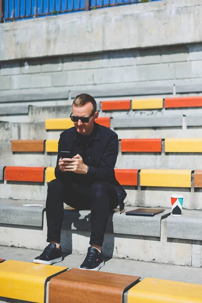 Şehirdeki şehir merkezinde dinlenirken gülümseyen modaya uygun giyinmiş neşeli hippi adam boş zamanlarında cep telefonu kullanıyor. Güneş gözlüklü mutlu Orta Doğulu erkek blogcu seyahat yaşam tarzının keyfini çıkarıyor.