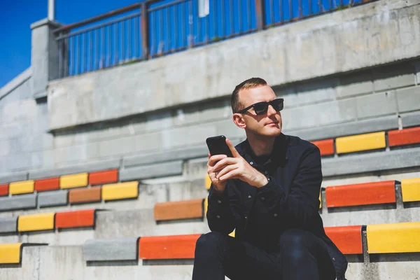 Şehirdeki şehir merkezinde dinlenirken gülümseyen modaya uygun giyinmiş neşeli hippi adam boş zamanlarında cep telefonu kullanıyor. Güneş gözlüklü mutlu Orta Doğulu erkek blogcu seyahat yaşam tarzının keyfini çıkarıyor.