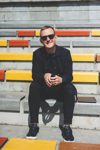 Şehirdeki şehir merkezinde dinlenirken gülümseyen modaya uygun giyinmiş neşeli hippi adam boş zamanlarında cep telefonu kullanıyor. Güneş gözlüklü mutlu Orta Doğulu erkek blogcu seyahat yaşam tarzının keyfini çıkarıyor.