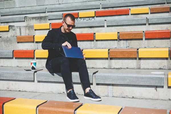 Genç işadamı ofisinin önündeki bankta otururken bilgisayarla çalışıyor.