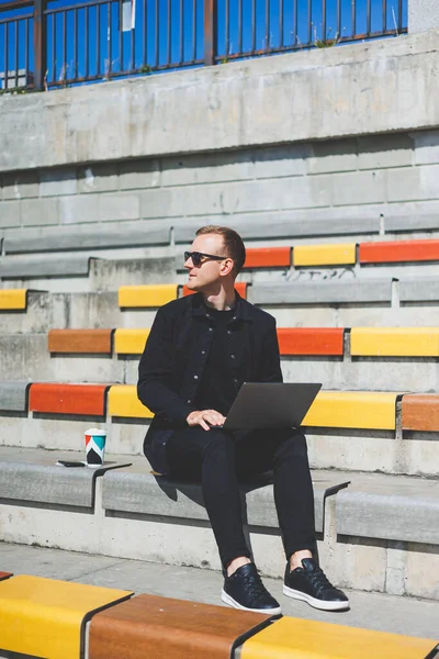 Genç işadamı ofisinin önündeki bankta otururken bilgisayarla çalışıyor.