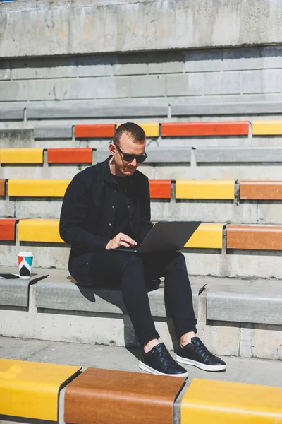 Genç işadamı ofisinin önündeki bankta otururken bilgisayarla çalışıyor.