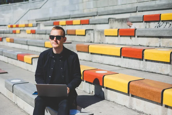 Genç işadamı ofisinin önündeki bankta otururken bilgisayarla çalışıyor.